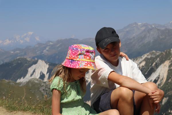 Two children are sitting on a mountain ridge, with a vast expanse of rugged mountains stretching into the distance under a clear blue sky